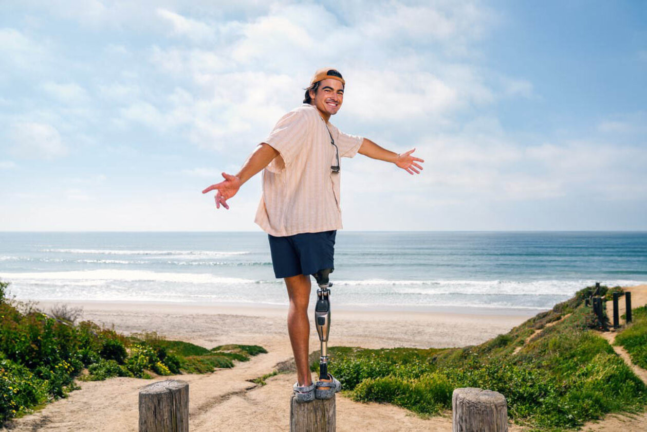 Knieprothesenträger Kaleb balanciert auf einem Holz-Pfahl in den Dünen. Die Sonne scheint, am Horizont sieht man das blaue Meer. Kaleb lacht. Er kann trotz Knieprothese die Balance halten.