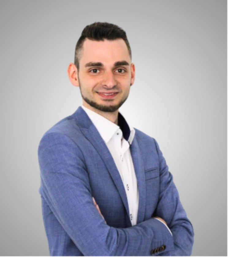 Ein lächelnder junger Mann mit dunklen Haaren und Dreitagebart. Er trägt ein weißes Hemd, darüber ein mittelblaues Sacko und hat die Arme verschränkt.