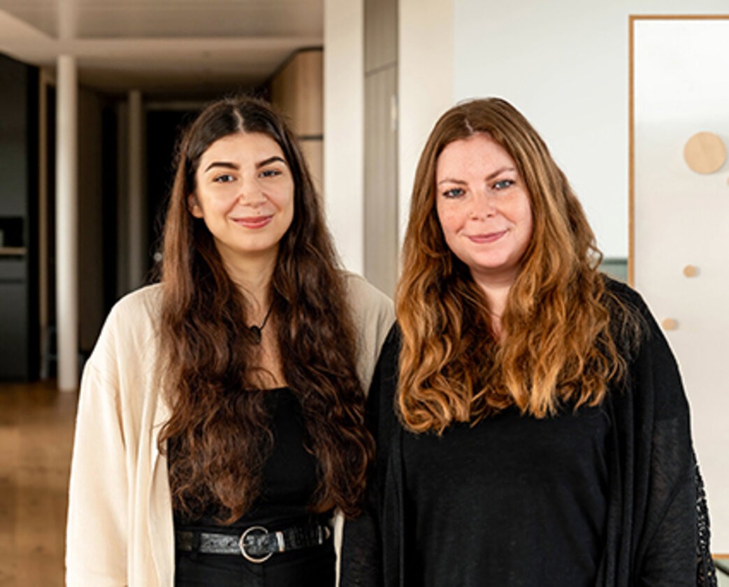 Zwei lächelnde, langhaarige Frauen stehen nebeneinander und lächeln in die Kamera.