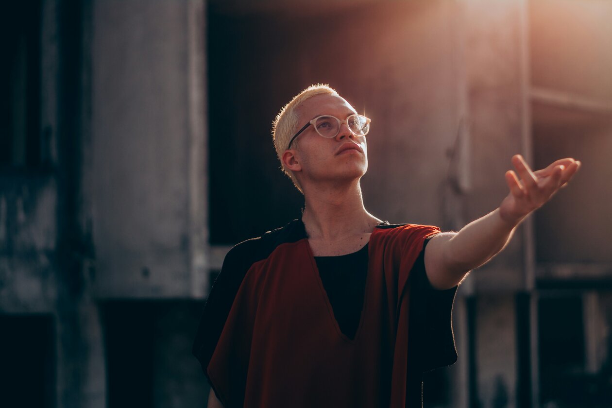 Ein Mann mit blondierten kurzen Haaren und dunkler Brille streckt die Hand in Richtung eines Lichtpunktes aus. 