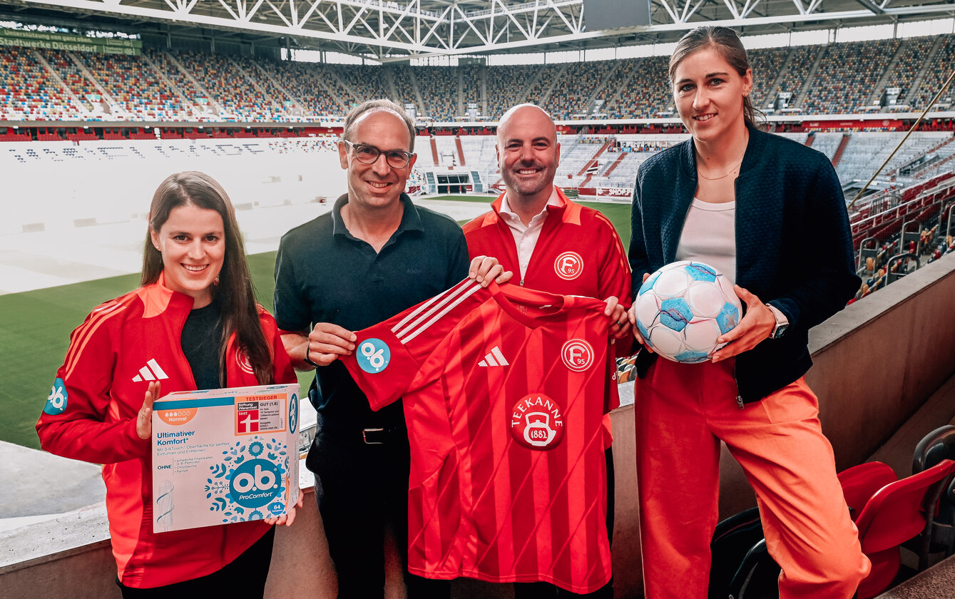 2 Füßballspielerinnen und 2 Männer halten ein rotes Trikot in die Kamera